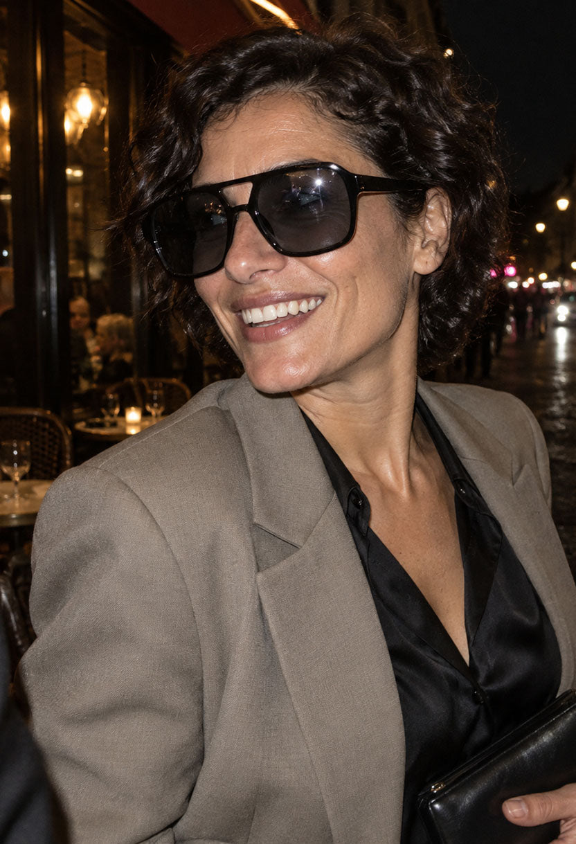 Smiling woman in a beige blazer wearing NEW STATEMENT AZTEC sunglasses on a lit street terrace at night #color_black