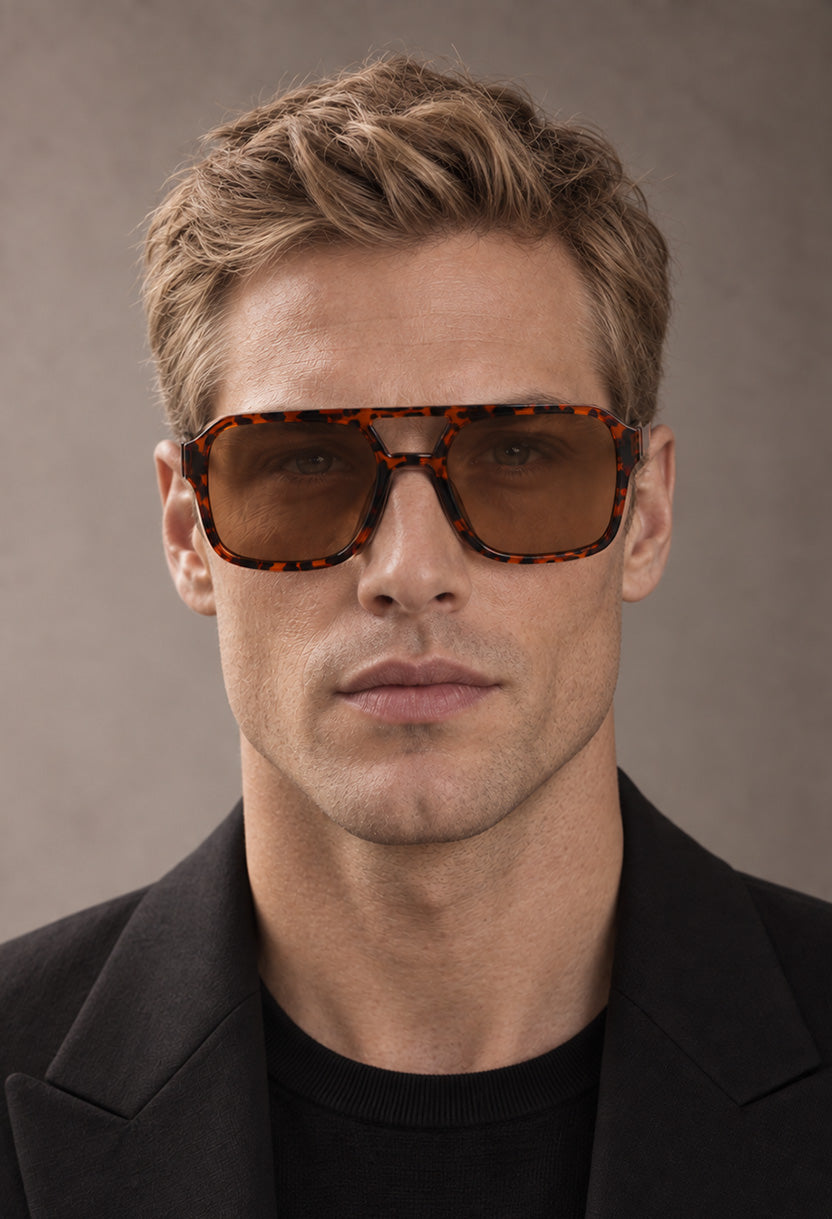 Smiling man in a navy polo wearing NEW STATEMENT AZTEC sunglasses by a columned facade at dusk #color_leopard