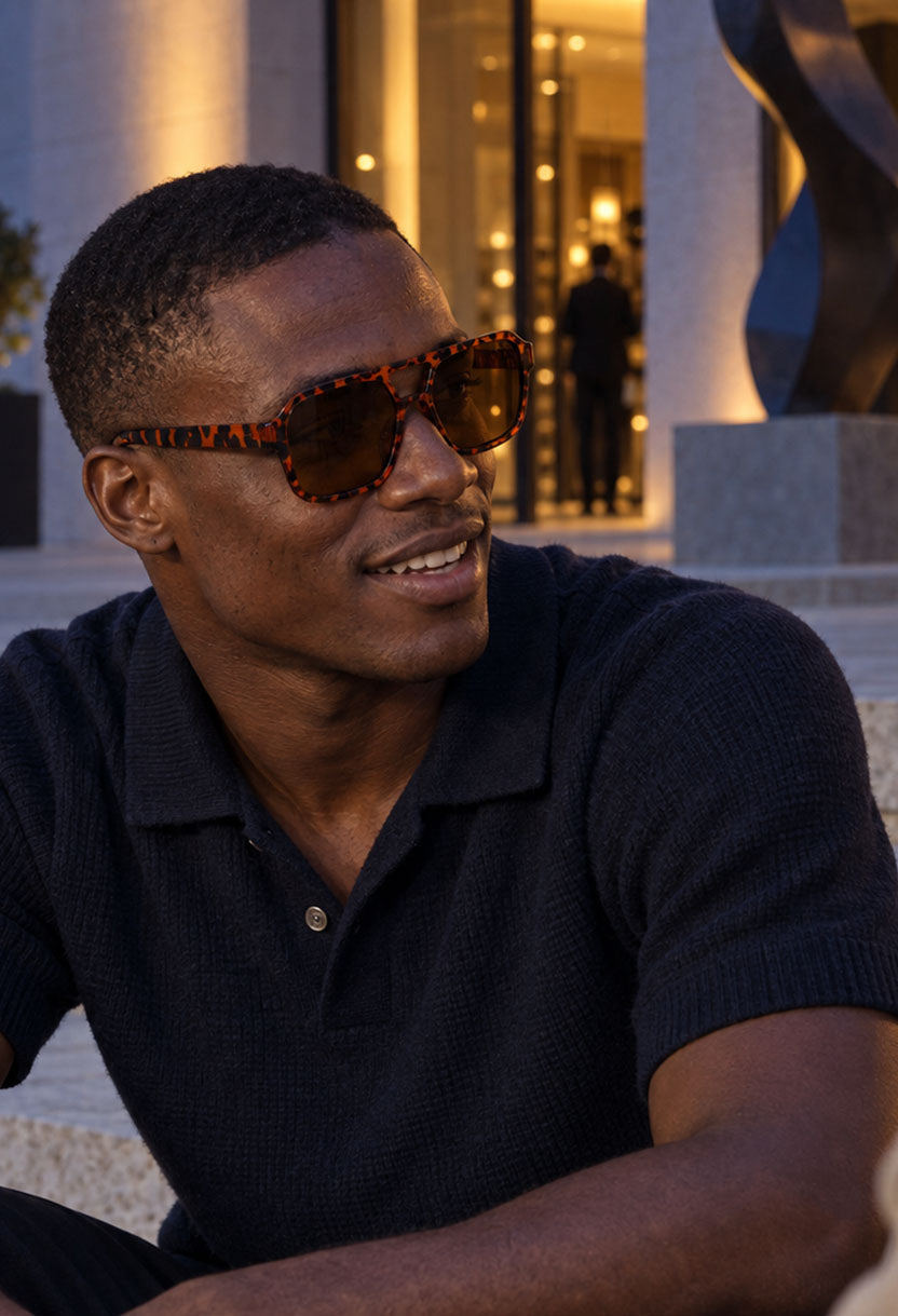 Smiling man in a navy polo wearing NEW STATEMENT AZTEC sunglasses by a columned facade at dusk #color_leopard