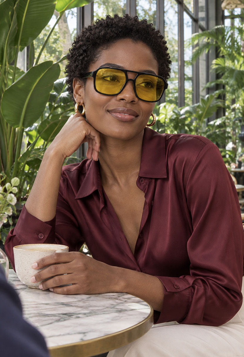 Woman in a burgundy silk blouse wearing NEW STATEMENT AZTEC sunglasses in a plant-filled conservatory #color_yellow