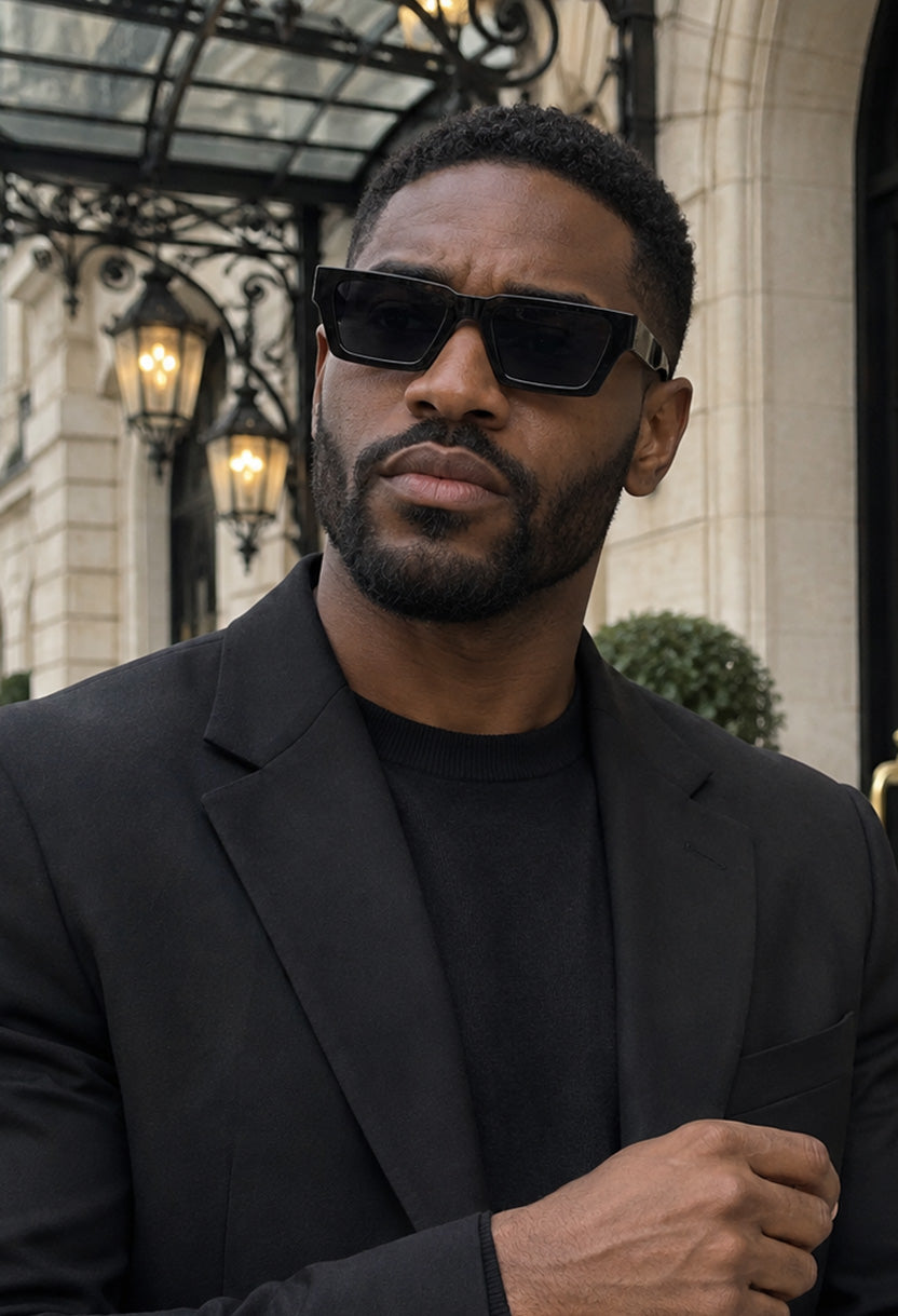 Man in a black blazer wearing NEW STATEMENT NEXUS sunglasses in black outside a grand hotel entrance #color_black